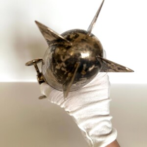 May include: A metallic, spherical object with multiple pointed projections. The object has a tarnished, bronze-like appearance and is held by a gloved hand. The design suggests a nautical or industrial theme.