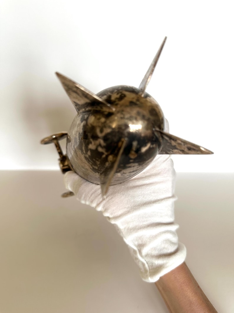 May include: A metallic, spherical object with multiple pointed projections. The object has a tarnished, bronze-like appearance and is held by a gloved hand. The design suggests a nautical or industrial theme.