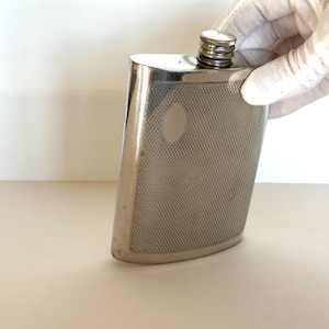 May include: A silver-toned metal flask with a curved, rectangular body and a screw-top cap. The front surface features a fine, cross-hatched diamond texture and a smooth, oval-shaped blank plaque area for engraving or personalization.