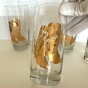 May include: Clear glass tumblers with a gold cowboy boot design. The boots have intricate white detailing. The glasses are cylindrical and appear to be empty. Several glasses are visible, with one being held by a gloved hand.