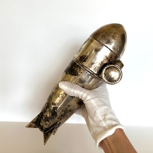 May include: A vintage silver cocktail shaker with a torpedo-like shape. The shaker has a rounded top, a central band, and a fin-like detail at the base. The metal has a tarnished appearance, suggesting age.