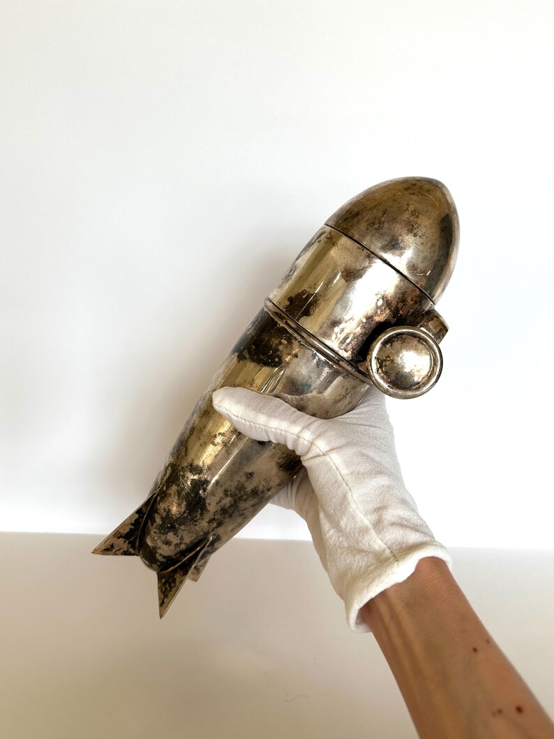 May include: A vintage silver cocktail shaker with a torpedo-like shape. The shaker has a rounded top, a central band, and a fin-like detail at the base. The metal has a tarnished appearance, suggesting age.