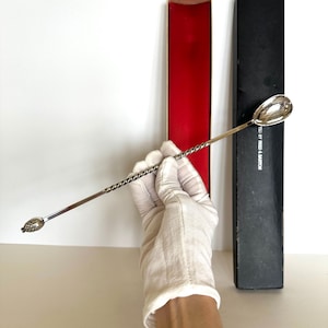 May include: A silver bar spoon with a twisted handle and a spoon-shaped bowl. The opposite end features a decorative design. The spoon is held near two rectangular boxes, one red and one black.
