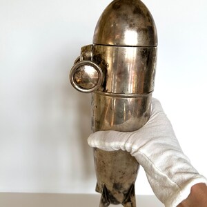 May include: A vintage silver-colored cocktail shaker with a rocket ship design. The shaker has a rounded top, a cylindrical body, and three fins at the base. A circular handle is attached to the side. The shaker is held by a gloved hand.