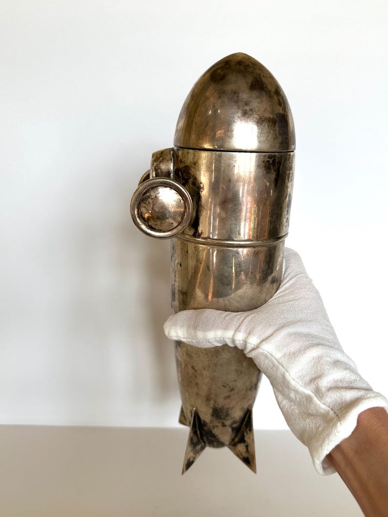 May include: A vintage silver-colored cocktail shaker with a rocket ship design. The shaker has a rounded top, a cylindrical body, and three fins at the base. A circular handle is attached to the side. The shaker is held by a gloved hand.
