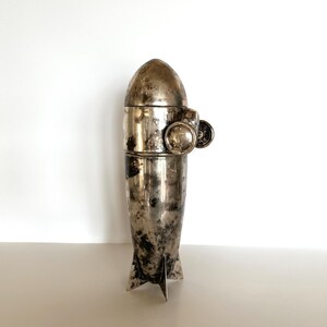 May include: A vintage silver-toned cocktail shaker with a rocket ship design. The shaker has a bullet-shaped top, a cylindrical midsection, and fins at the base. It features a small, round handle and a decorative element on the side.