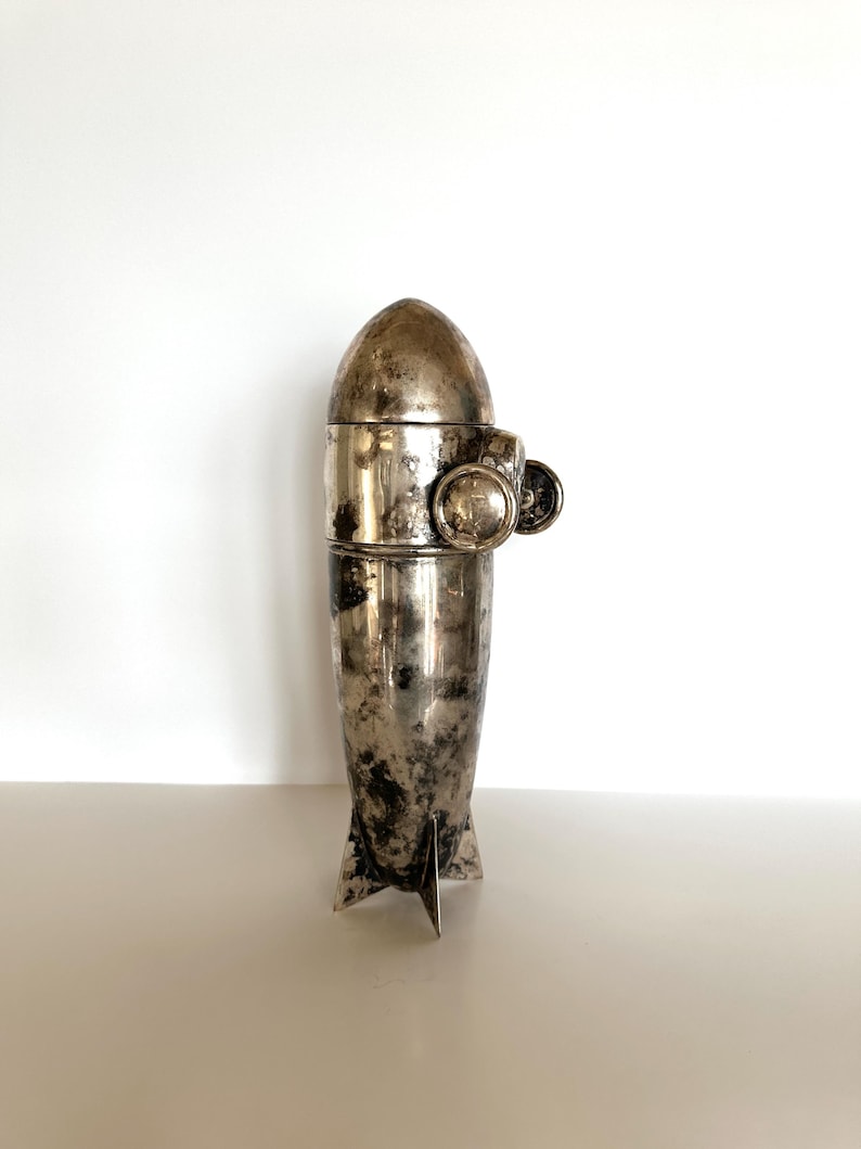 May include: A vintage silver-toned cocktail shaker with a rocket ship design. The shaker has a bullet-shaped top, a cylindrical midsection, and fins at the base. It features a small, round handle and a decorative element on the side.