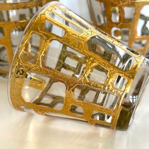 May include: Three clear glass tumblers featuring a decorative gold-colored grid pattern with a textured, crackled finish. The glasses have a cylindrical shape with a wide opening and a smooth, transparent base, showcasing a repeating geometric design.