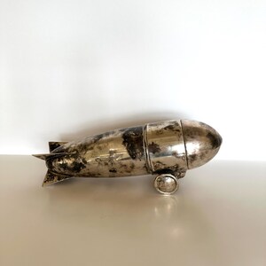 May include: A vintage silver-toned metal zeppelin-shaped object with a tarnished finish. The object has a cylindrical body with rounded ends, small wheels, and tail fins. It appears to be a decorative item or container.
