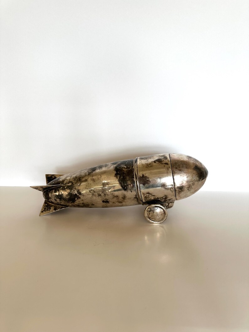 May include: A vintage silver-toned metal zeppelin-shaped object with a tarnished finish. The object has a cylindrical body with rounded ends, small wheels, and tail fins. It appears to be a decorative item or container.