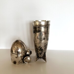 May include: Two antique silver-toned items. One is a tall, cylindrical cocktail shaker with a flared top and three fins at the base. The other is a small, bullet-shaped container with a lid and a small handle.