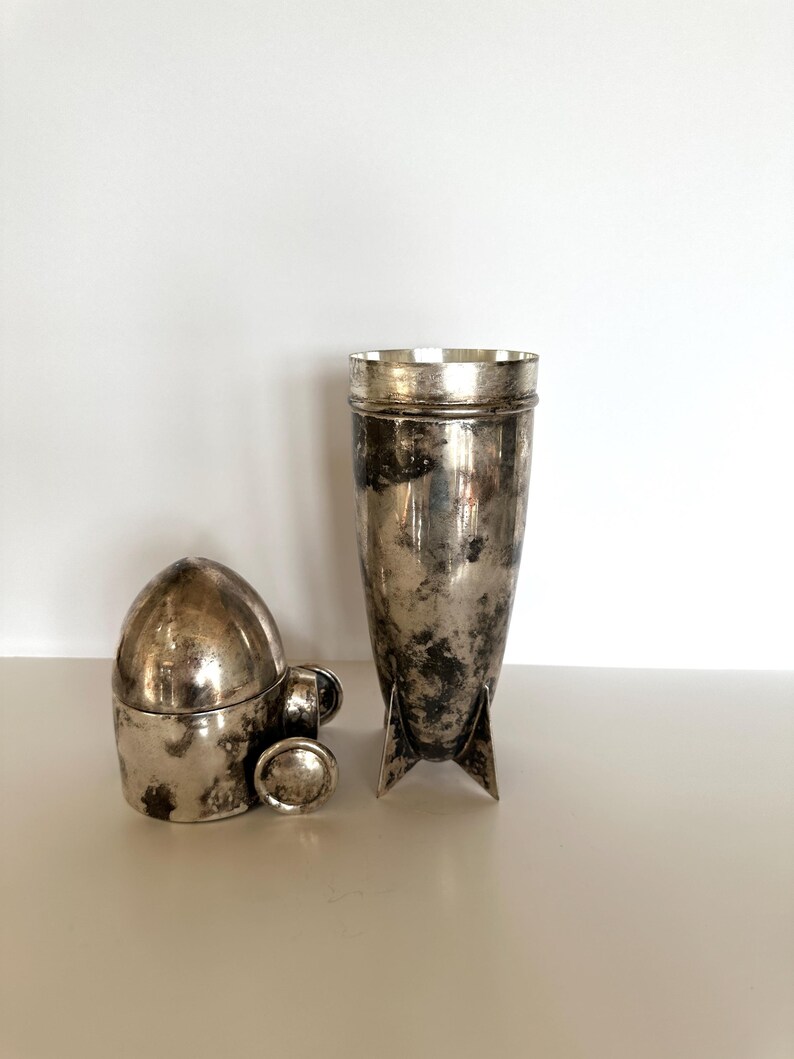 May include: Two antique silver-toned items. One is a tall, cylindrical cocktail shaker with a flared top and three fins at the base. The other is a small, bullet-shaped container with a lid and a small handle.