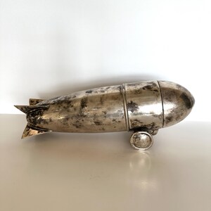 May include: A silver-colored blimp-shaped object with a textured surface. The blimp has a rounded front and rear, with small fins at the back. It rests on two small, circular wheels. The object appears to be made of metal.