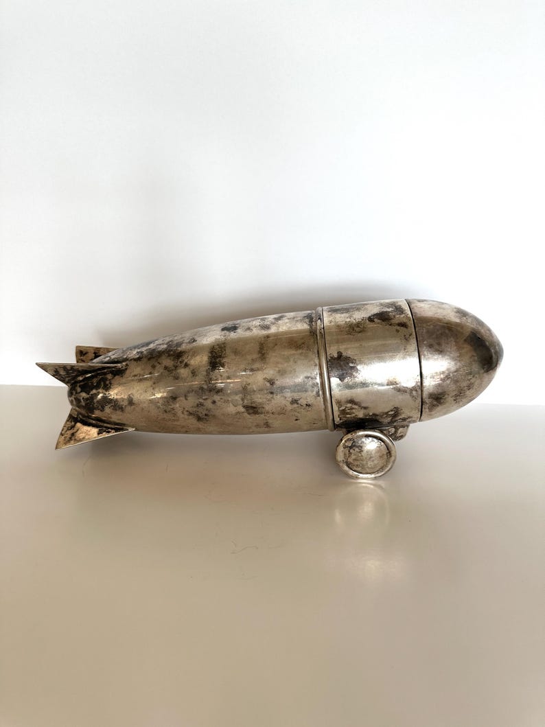 May include: A silver-colored blimp-shaped object with a textured surface. The blimp has a rounded front and rear, with small fins at the back. It rests on two small, circular wheels. The object appears to be made of metal.