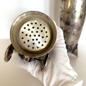 May include: Vintage silver cocktail shaker with a strainer and a separate conical container. The strainer has a circular design with multiple small holes. The metal shows signs of age and use, with a slightly tarnished appearance.