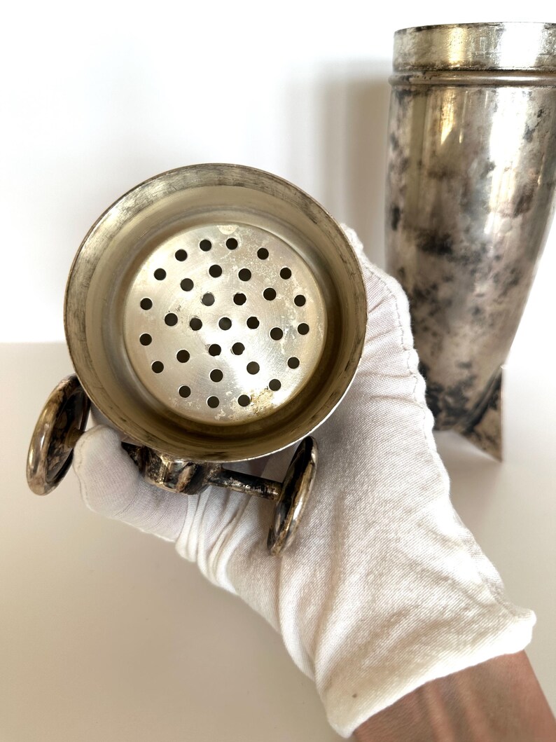 May include: Vintage silver cocktail shaker with a strainer and a separate conical container. The strainer has a circular design with multiple small holes. The metal shows signs of age and use, with a slightly tarnished appearance.