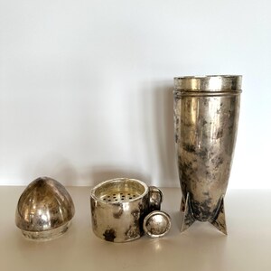 May include: A vintage silver cocktail shaker set, disassembled. The set includes a shaker, a strainer with a small handle, a cap, and a lid. The pieces have a tarnished, aged appearance.