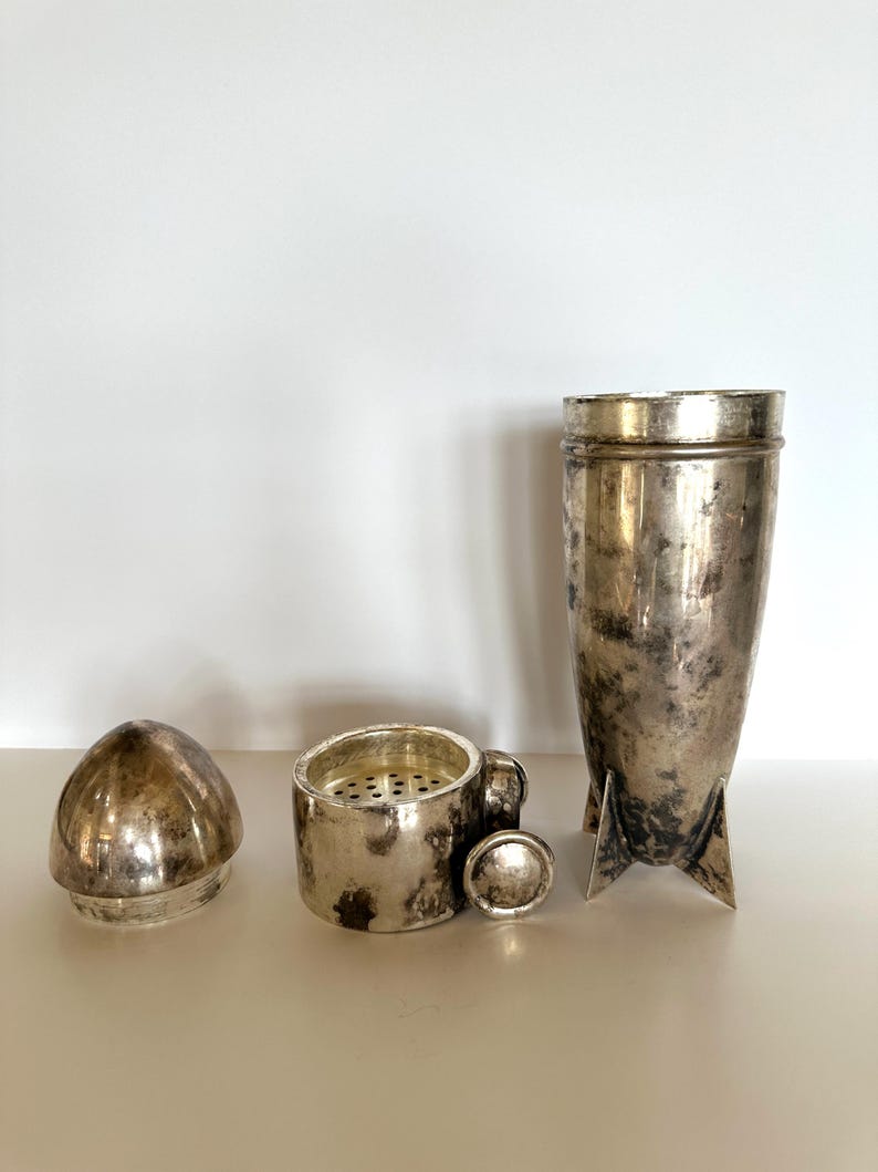 May include: A vintage silver cocktail shaker set, disassembled. The set includes a shaker, a strainer with a small handle, a cap, and a lid. The pieces have a tarnished, aged appearance.