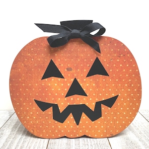 May include: Orange and black Halloween pumpkin with a black bow and a jack-o'-lantern face. The pumpkin is decorated with white polka dots.