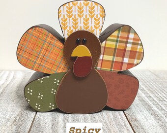 May include: A wooden turkey figurine with a brown body, a red beak, and black eyes. The turkey's feathers are made of patterned fabric in shades of orange, green, and brown. The word "Spicy" is written on a white tag below the turkey.