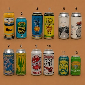 May include: A collection of twelve beer cans with various designs and labels. The cans feature different colors, graphics, and text, including "Madonna," "Cone Head," "Little Sip," and "Pale Ale." The cans are arranged on a brown surface.