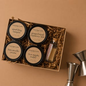 May include: A gift set featuring four scented soy candles in black tins with labels reading "sparkling sangria", "gingerbread espresso 'tini", "limoncello creme", and "hot maple toddy". A small glass bottle of matches and two stainless steel jiggers are also included.