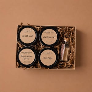 May include: A gift set containing four black candle tins, each labelled with a different name: "north end", "dunkin run", "boston tea party", and "the cape". A small glass bottle of matches is included. The candles are hand-poured soy candles.