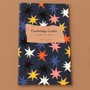 May include: A notebook with a navy blue cover patterned with colourful starbursts in white, yellow, orange, pink, and blue. The cover has a brown label with the text "Cambridge Crafter scrapbook paper girly pop."