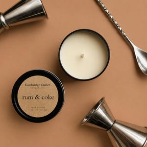 May include: Overhead shot of two silver jiggers, a silver bar spoon, and two black candle tins on a tan surface. One candle tin is open, revealing a white candle. The other tin reads "Rum & Coke".