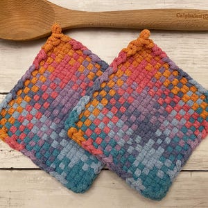May include: Two square, woven potholders in a gradient of colors, including pink, orange, purple, and blue. A wooden spoon rests above the potholders. The potholders are handmade and have a textured appearance, suitable for kitchen use.
