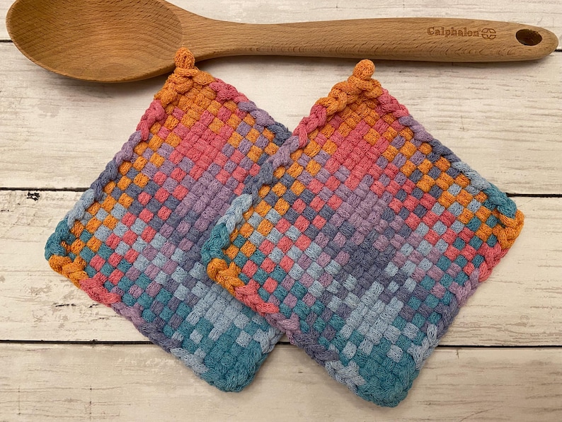 May include: Two square, woven potholders in a gradient of colors, including pink, orange, purple, and blue. A wooden spoon rests above the potholders. The potholders are handmade and have a textured appearance, suitable for kitchen use.