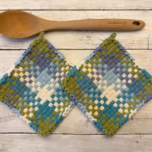 May include: Two square potholders featuring a woven checkerboard pattern in shades of olive green, light blue, and cream, positioned below a wooden spoon with the brand name Calphalon engraved on the handle near the circular hanging hole.