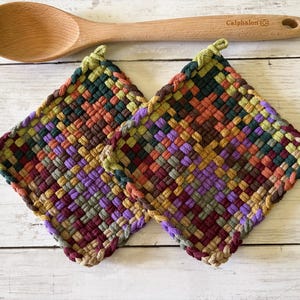 May include: Two square potholders featuring a woven pattern in shades of purple, green, orange, and dark red, positioned below a light-colored wooden spoon with the brand name Calphalon engraved on the handle.