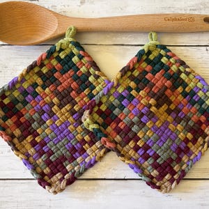 May include: Two square potholders featuring a woven pattern in shades of purple, green, orange, and brown, each with a small loop at the top corner. A light-colored wooden spoon with the brand name Calphalon engraved on the handle is positioned above.