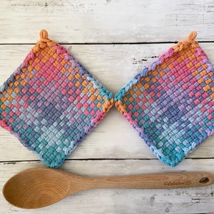 May include: Two square, woven potholders in a gradient of colors including pink, orange, blue, and purple. A wooden spoon rests below the potholders. The potholders have a loop for hanging.