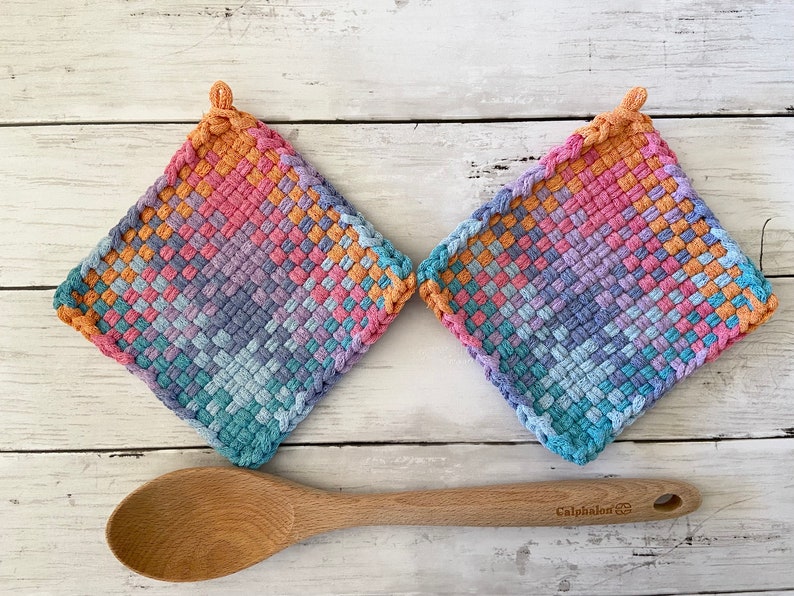 May include: Two square, woven potholders in a gradient of colors including pink, orange, blue, and purple. A wooden spoon rests below the potholders. The potholders have a loop for hanging.