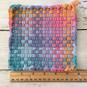 May include: A square, woven potholder in a gradient of colors, including pink, orange, blue, and purple. The potholder is approximately 6 inches wide and 6 inches long. It has a loop for hanging and is made of a tightly woven material.