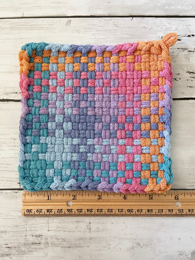 May include: A square, woven potholder in a gradient of colors, including pink, orange, blue, and purple. The potholder is approximately 6 inches wide and 6 inches long. It has a loop for hanging and is made of a tightly woven material.