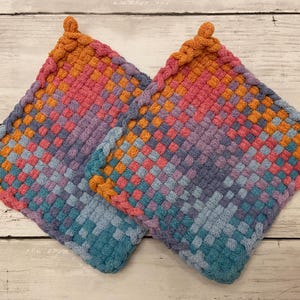 May include: Two woven potholders in a square shape. The potholders feature a colorful gradient pattern, transitioning from orange to pink, purple, and blue. Each potholder has a braided edge and a loop for hanging.