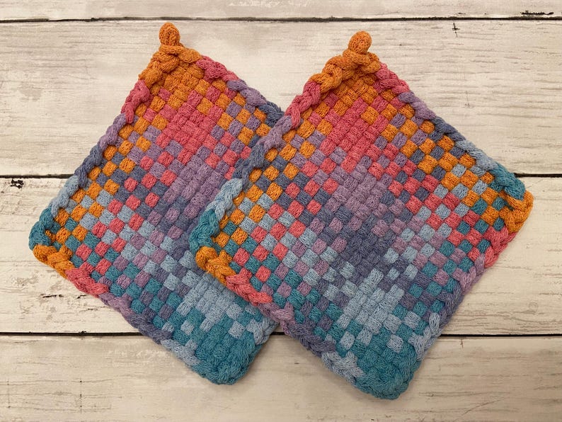 May include: Two woven potholders in a square shape. The potholders feature a colorful gradient pattern, transitioning from orange to pink, purple, and blue. Each potholder has a braided edge and a loop for hanging.