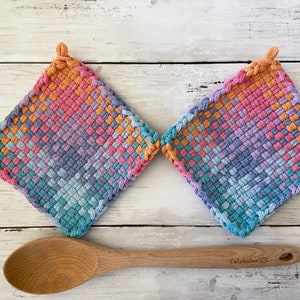 May include: Two square, woven potholders in a gradient of colors, including orange, pink, purple, and blue. A wooden spoon rests below the potholders. The potholders have a loop for hanging.