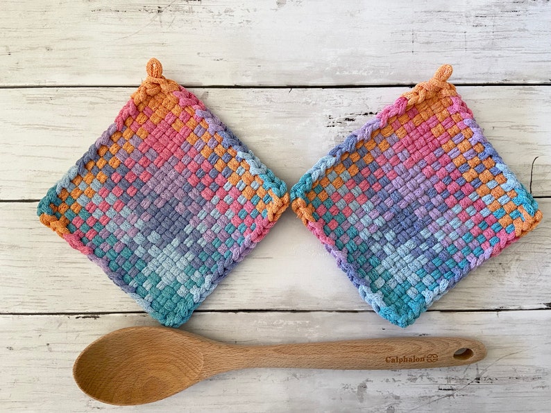 May include: Two square, woven potholders in a gradient of colors, including orange, pink, purple, and blue. A wooden spoon rests below the potholders. The potholders have a loop for hanging.