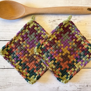 May include: Two square potholders featuring a woven pattern in shades of purple, green, orange, and dark teal, each with a small loop at the top corner. A light-colored wooden spoon with the brand name Calphalon engraved on the handle is included.