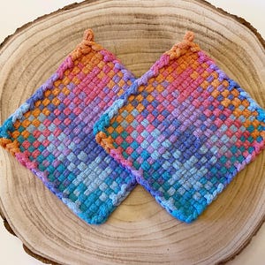 May include: Two square, woven potholders in a vibrant array of colors, including pink, orange, blue, and purple. The potholders are placed on a wooden surface, showcasing their handmade texture and design. Each potholder has a small loop for hanging.