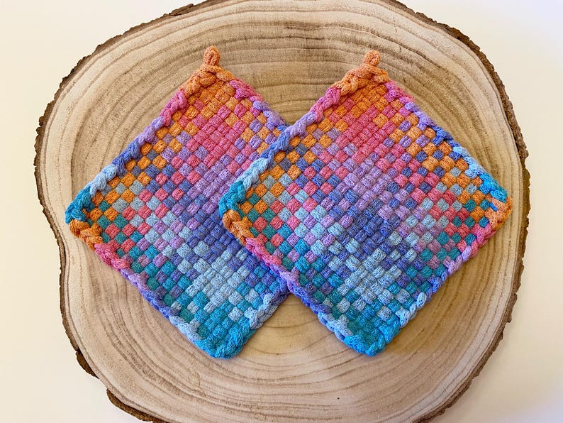 May include: Two square, woven potholders in a vibrant array of colors, including pink, orange, blue, and purple. The potholders are placed on a wooden surface, showcasing their handmade texture and design. Each potholder has a small loop for hanging.