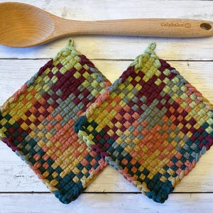 May include: Two square woven potholders featuring a multicolored checkered pattern in shades of burgundy, olive green, mustard yellow, and teal, accompanied by a light-colored wooden spoon with the brand name Calphalon engraved on the handle.