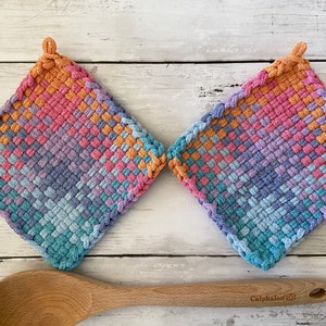 May include: Two square, woven potholders in a diamond pattern, featuring a gradient of colors including orange, pink, purple, and blue. Each potholder has a loop for hanging. A wooden spoon rests below.