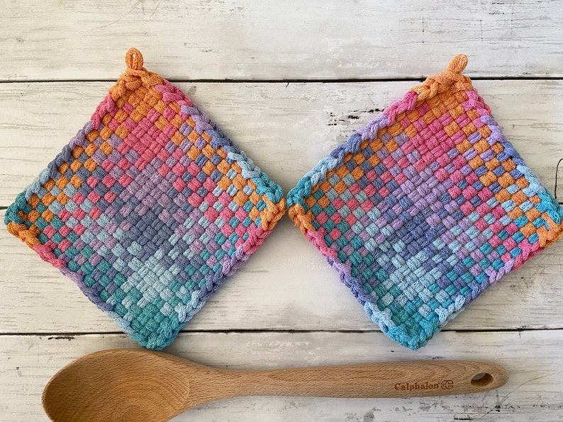 May include: Two square, woven potholders in a diamond pattern, featuring a gradient of colors including orange, pink, purple, and blue. Each potholder has a loop for hanging. A wooden spoon rests below.