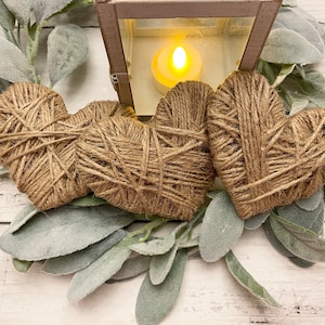 May include: Three brown twine hearts with a rustic, farmhouse style. The hearts are arranged in a semi-circle around a small lantern with a lit candle inside.