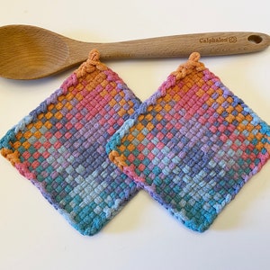 May include: Two square, woven potholders in shades of blue, pink, orange, and purple. A wooden spoon rests above them. The potholders have a braided edge and a loop for hanging. The spoon is light brown and has the word "Calphalon" printed on it.